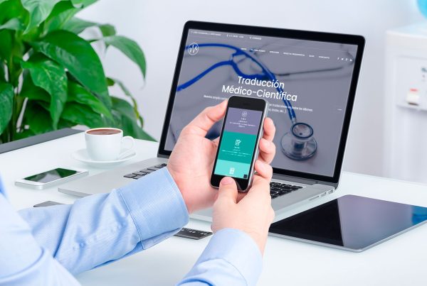 Mano sujetando un móvil que enseña el sitio web de traducción, con laptop detrás enseñando también el sitio web, ambos elementos en una oficina.